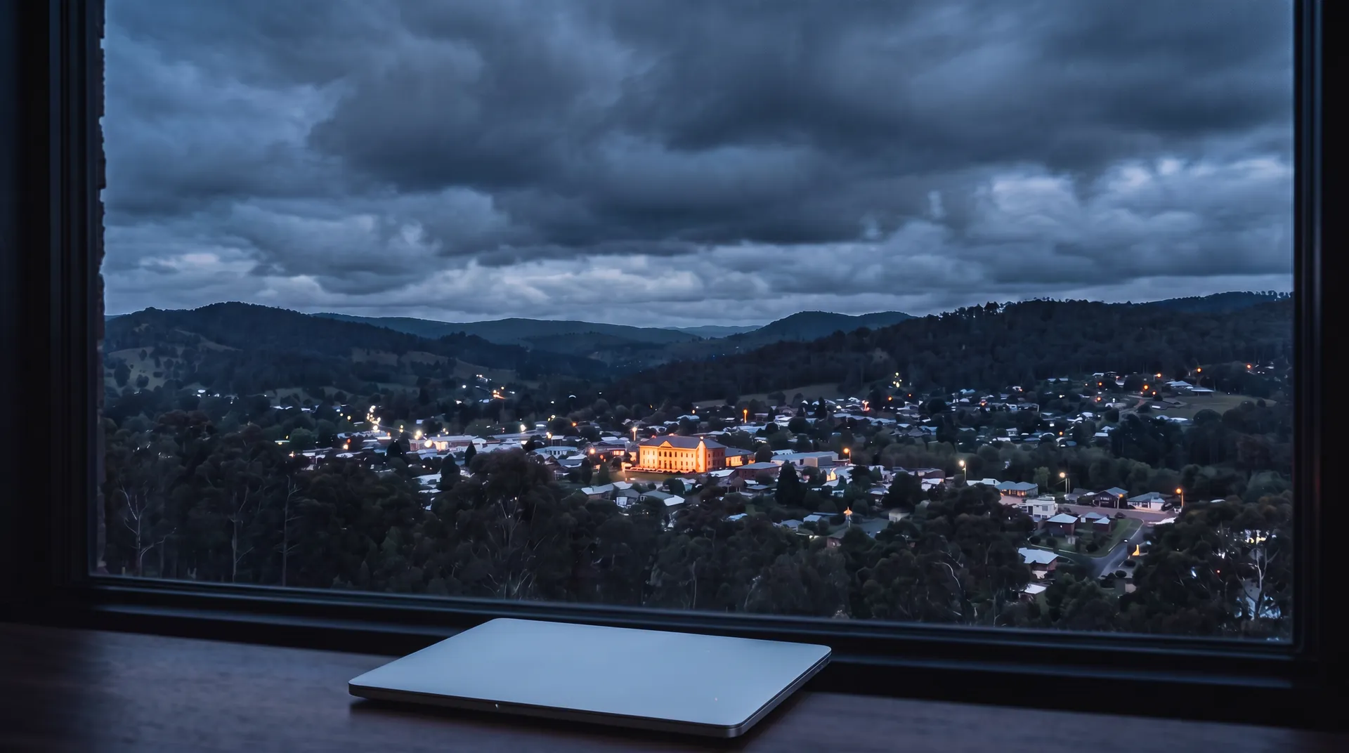 AI image: View from a window across a valley at blue hour, one building glows brighter than all the others, a closed laptop lies on the windowsill